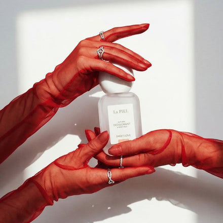 High-fashion shot of hands in sheer red gloves holding a bottle of La PIEL 'SWEET LOVE' Natural Deodorant against white background.